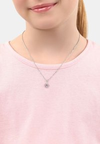 Silverfärgad halskedja med ett hjärtformat hänge som har en rosa ädelsten omgiven av små klara stenar, bärs över en rosa skjorta.