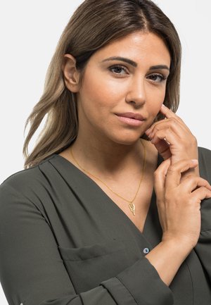 Ein anhängerförmiges Blatt aus Gold auf einer dünnen Kette, getragen über einer dunkelgrünen Bluse mit V-Ausschnitt und langen Ärmeln. Glatte, glänzende Texturen.