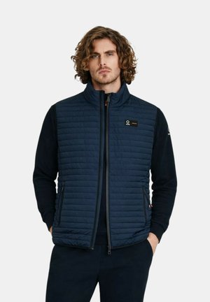 Uomo con capelli ricci che indossa un gilet trapuntato senza maniche blu navy sopra una camicia nera a maniche lunghe, in piedi con le mani nelle tasche.
