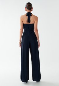 Mono de tirantes azul marino con un cuerpo ajustado, pantalones de pierna ancha y un lazo en el cuello. Textura suave, cierre de cremallera en la parte posterior.