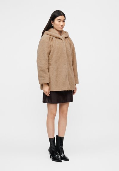 Μπεζ χνουδωτό υπερμεγέθες παλτό με κουκούλα, μπροστινές τσέπες, μακριά μανίκια, φορεμένο πάνω από καφέ δερμάτινη μίνι φούστα και μαύρες μποτάκια μέχρι τον αστράγαλο.