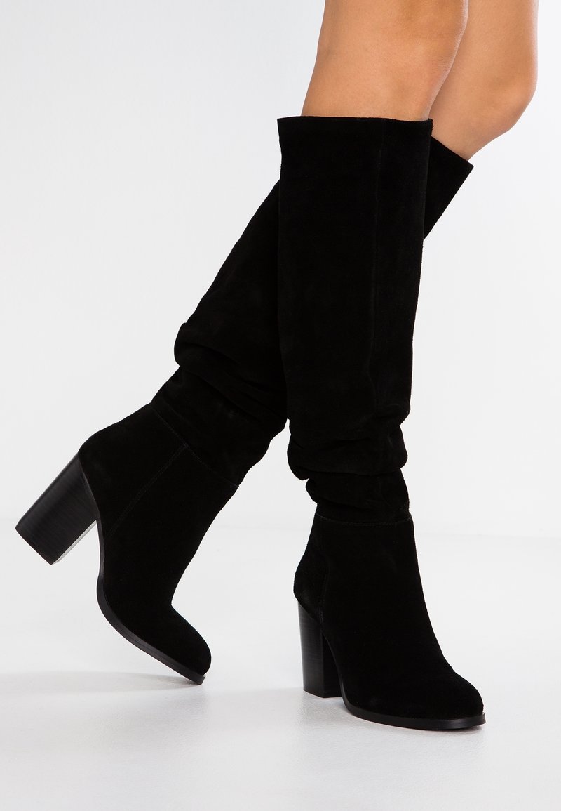 Bottes hautes en daim noir au-dessus du genou avec un design ample. Bout carré, talon épais, et texture lisse, idéales pour diverses tenues.