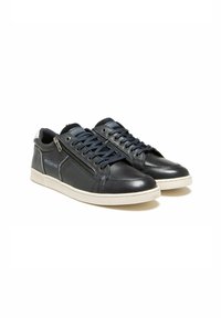 Une paire de baskets en cuir noir avec des zips sur le côté, des lacets navy et des semelles blanches, présentée sur un fond blanc.