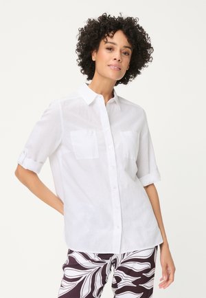 Donna con capelli ricci che indossa una camicia bianca abbottonata con maniche risvoltate e pantaloni a fantasia neri e bianchi, in piedi con una mano sui fianchi.