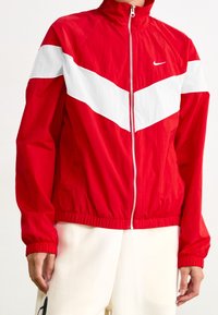 Chaqueta de chándal Nike de color rojo y blanco con cremallera, con puños y dobladillo elásticos, combinada con pantalones de color claro, vista desde el medio del pecho hasta las caderas.