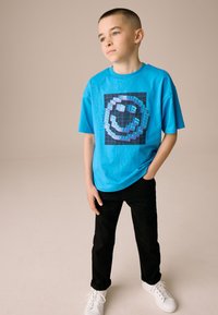 Turquoise T-shirt met korte mouwen en een gepixelde smiley-afbeelding, gecombineerd met zwarte broek en witte sneakers. Zachte stoftextuur.