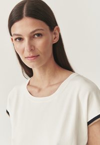 T-shirt bianco in cotone con colletto rotondo e maniche corte, con dettagli neri e beige sui bordi delle maniche. Texture liscia.