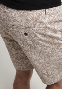 Shorts beiges avec un motif paisley blanc, fabriqués en tissu léger. Comprend une poche avant avec un bouton noir et un design à coupe relaxée.