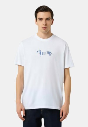 Modello maschile che indossa una T-shirt bianca con una grafica di un bassotto a righe blu, abbinata a pantaloni scuri, su uno sfondo chiaro e uniforme.