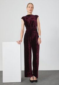 Combinaison en velours bordeaux avec un col haut et des manches courtes, présentant un design de taille torsadée et une coupe large, assortie à des talons noirs.