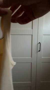 Krémově barevná žebrovaná látka držena vzhůru, ukazující měkkou texturu a zakřivené okraje, s rozmazanou bílou skříní v pozadí.