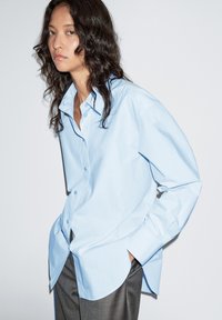 Lichtblauw katoenen overhemd met knoopsluiting, voorzien van een klassieke kraag, lange mouwen en een afgeronde zoom. Gecombineerd met grijze broek.