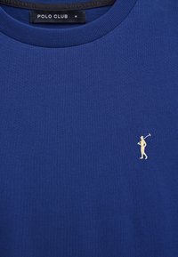 T-shirt bleu à col ras du cou avec un petit logo de joueur de polo beige sur la poitrine et une étiquette noire portant l'inscription "POLO CLUB M" à l'intérieur du col.