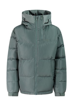 Jong meisje met lang krullend haar glimlachend, draagt een teal donzen jas, zwarte broek en zwarte schoenen, staat met handen in de zakken.