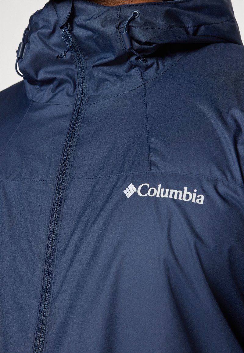Dunkelblaues Columbia-Jackett mit hohem Kragen, vollem Reißverschluss und verstellbaren Zugbändern. Mit dem weißen Columbia-Logo, das prominent dargestellt ist.