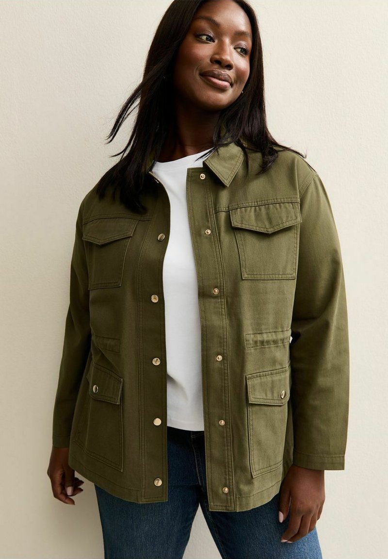 Vrouw draagt een olijfgroene utility-jas met knopen en klepzakken over een wit shirt en blauwe spijkerbroek, staand tegen een neutrale achtergrond.