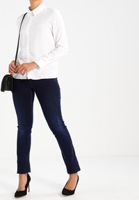 Donna che indossa una camicia bianca con bottoni, jeans blu scuri, tacchi alti neri e porta una piccola borsa nera a tracolla.