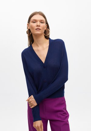 Tmavě modrý cardigan z měkké tkaniny, s véčkovým výstřihem, dlouhými rukávy a dvěma fialovými knoflíky vpředu, který je kombinován s fialov�ými kalhotami.