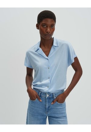 Vrouw met kort haar, gekleed in een lichtblauw kort mouw knoopjesoverhemd en blauwe spijkerbroek, staand met handen in de zakken tegen een effen achtergrond.