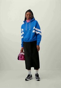 Blå hettegenser med hvite striper, svarte vide bukser, rosa håndveske og sneakers. Har snor i hetta og en rosa genser med høy hals under.