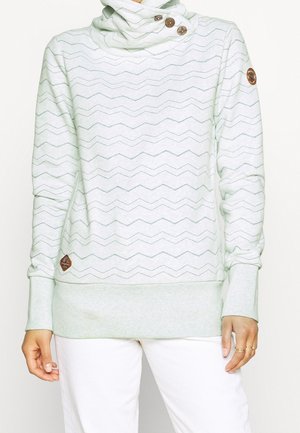 Vrouw draagt lichtgrijze pullover hoodie met zigzagpatroon, drie houten knopen bij de hals en leren merkpatches op mouw en zoom.