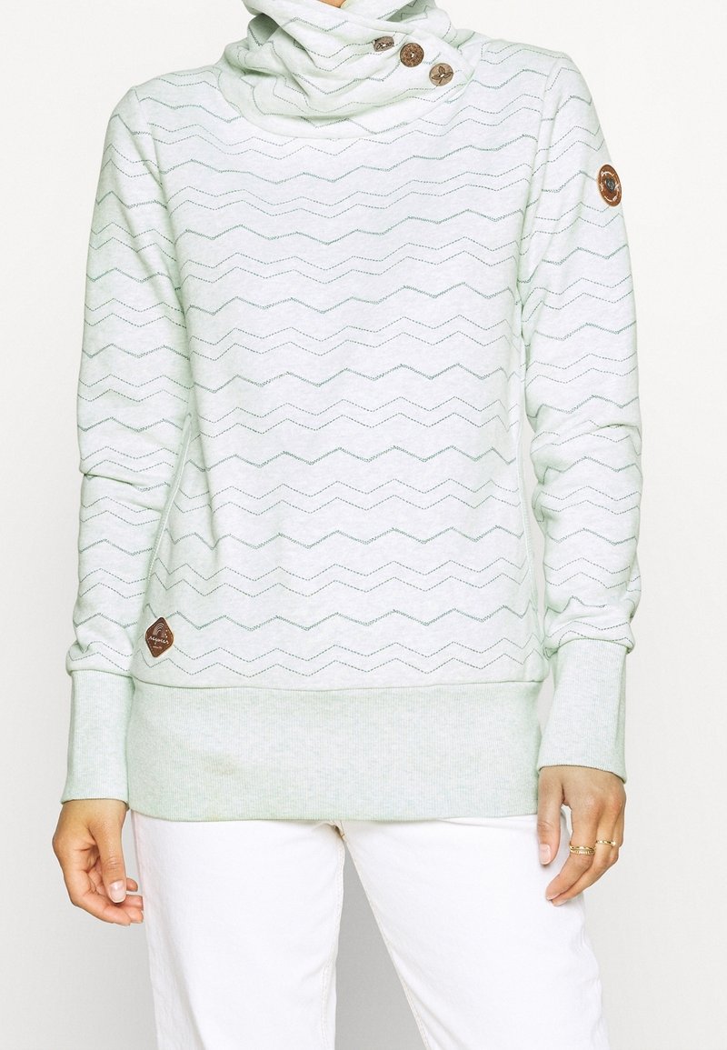 Femme portant un sweat à capuche gris clair avec motif en zigzag, trois boutons en bois près du cou, et des patchs en cuir de la marque sur la manche et l'ourlet.