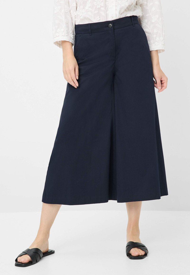 Navy wide-leg trousers made of smooth fabric, featuring a front button closure and side pockets. Worn with black slide sandals.