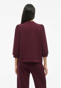 Burgunderfarget genser med trekvartlange ermer, ribbestrikket nederkant og mykt stoff. Har en avslappet passform og skulderplisseringer for ekstra volum.