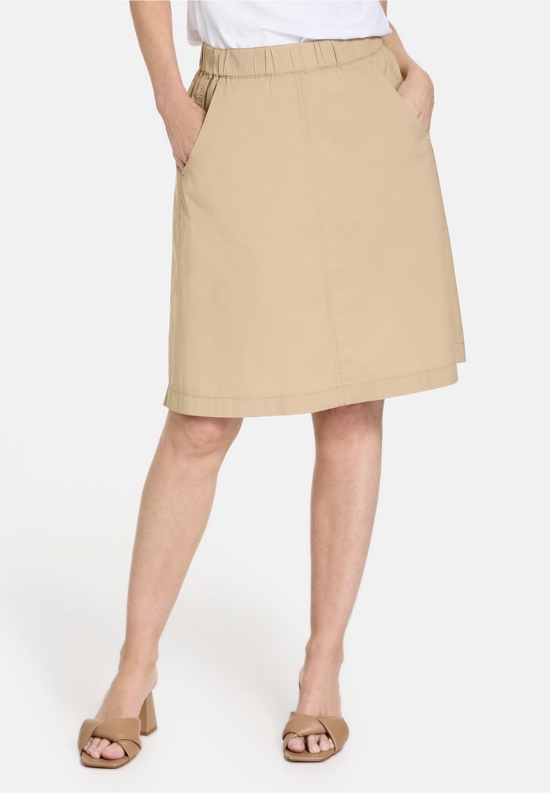 Jupe kaki jusqu'aux genoux en tissu léger. Elle possède une taille élastique et des poches latérales. Portée avec des sandales à talons blocs couleur beige.