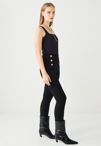 Schwarzer ärmelloser Body kombiniert mit hoch taillierten schwarzen Hosen, die vier goldene Knöpfe haben, und schwarzen Ankle Boots mit spitzer Schuhspitze.