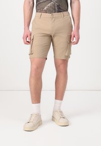 Shorts cargo beiges avec poches latérales, fabriqués dans un tissu extensible, dotés d'une fermeture à bouton et d'un design à l'avant plat. Portés avec des chaussettes blanches et des chaussures.