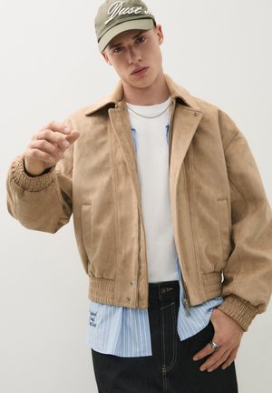 Unge mand iført en beige jakke over en stribet skjorte og hvid t-shirt, olivengrøn kasket, sølvkædehalskæde og en ring på sin højre hånd.
