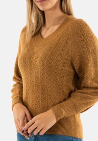 Suéter de cuello en V y manga larga en color mostaza con un patrón de chevron texturizado. Puños y dobladillo acanalados. Combinado con jeans azules.