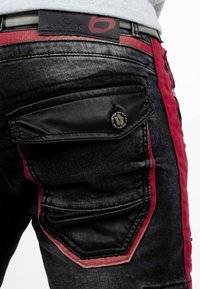 Jeans en denim noir avec des accents rouges, présentant une poche arrière avec un rabat texturé noir et un détail de bouton en métal, ainsi qu'une ceinture de marque.