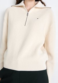 Kobieta nosząca kremowy sweter z prążkowanej dzianiny z półzamkiem oraz małym haftowanym logo na piersi i czarne spodnie.