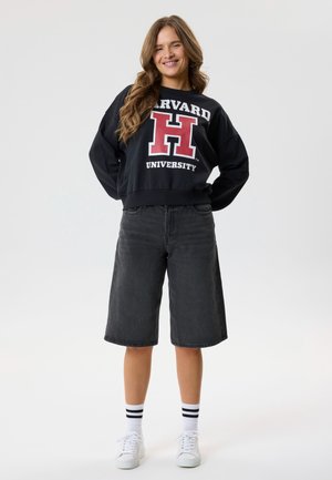Kvinde står og smiler, iført en sort Harvard University sweatshirt, sorte knælange shorts, hvide sokker med sorte striber og hvide sneakers.