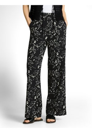 Kobieta nosząca czarne spodnie z szerokimi nogawkami w biały kwiatowy wzór, ręce w kieszeniach, w zestawieniu z czarnymi sandałami na płaskiej podeszwie.