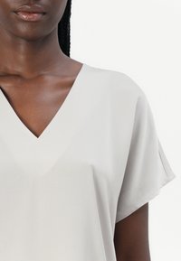 Záber zblízka na osobu v svetlosivom tričku s krátkym rukávom a výstrihom do V na obyčajnom bielom pozadí.