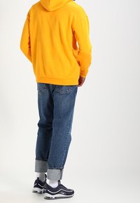 Felpa con cappuccio gialla, jeans blu con orli risvoltati e sneakers navy con accenti bianchi. Presenta una vestibilità comoda e una texture di tessuto standard.