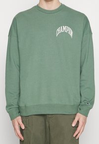 Zelená bavlněná mikina se žebrovanými manžetami a lemem. Na hrudi se nachází zakřivené logo "CHAMPION" v bílé barvě. Volný střih.