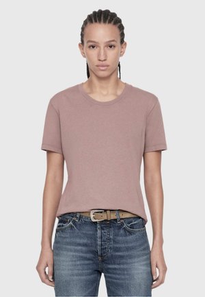 Person with braided hair wearing a dusty pink short-sleeve t-shirt tucked into blue jeans with a brown belt on a plain light background.