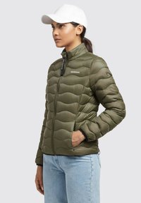 Olivovo zelená puff jacket s predným zipsom, zvlnenou prešívanou textúrou, stojatým golierom a dvoma bočnými vreckami; spárovaná so svetlo modrými džínsami.