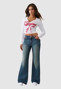 Witte cropped top met lange mouwen en een roze grafische print, gecombineerd met blauwe flared jeans met vervaagde details. Model draagt witte sneakers.