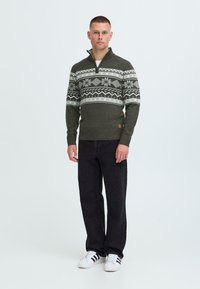 Pulover tricotat de culoare verde închis, cu un model geometric alb, guler înalt cu fermoar la jumătate; asortat cu pantaloni negri și adidași albi.