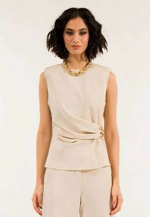 Top beige senza maniche con design attorcigliato in vita, caratterizzato da un dettaglio ad anello dorato. Texture liscia e taglio semplice adatto per diverse occasioni.