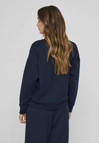 Tumši zila svītrainā piedurkņu krekls ar nolaižamām plecu līnijām un garām piedurknēm, izgatavots no mīksta materiāla, kas nodrošina brīvu piegriezumu un ribotas aproces.