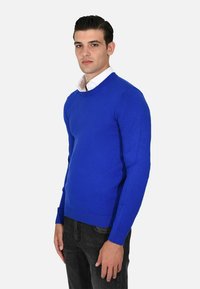 Maglione blu lavorato a maglia con scollatura rotonda e polsini a coste, indossato sopra una camicia bianca con colletto, abbinato a jeans scuri.