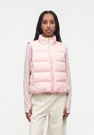 Donna con i capelli intrecciati che indossa un piumino rosa chiaro sopra una camicia a maniche lunghe e pantaloni color crema, in piedi davanti a uno sfondo semplice.