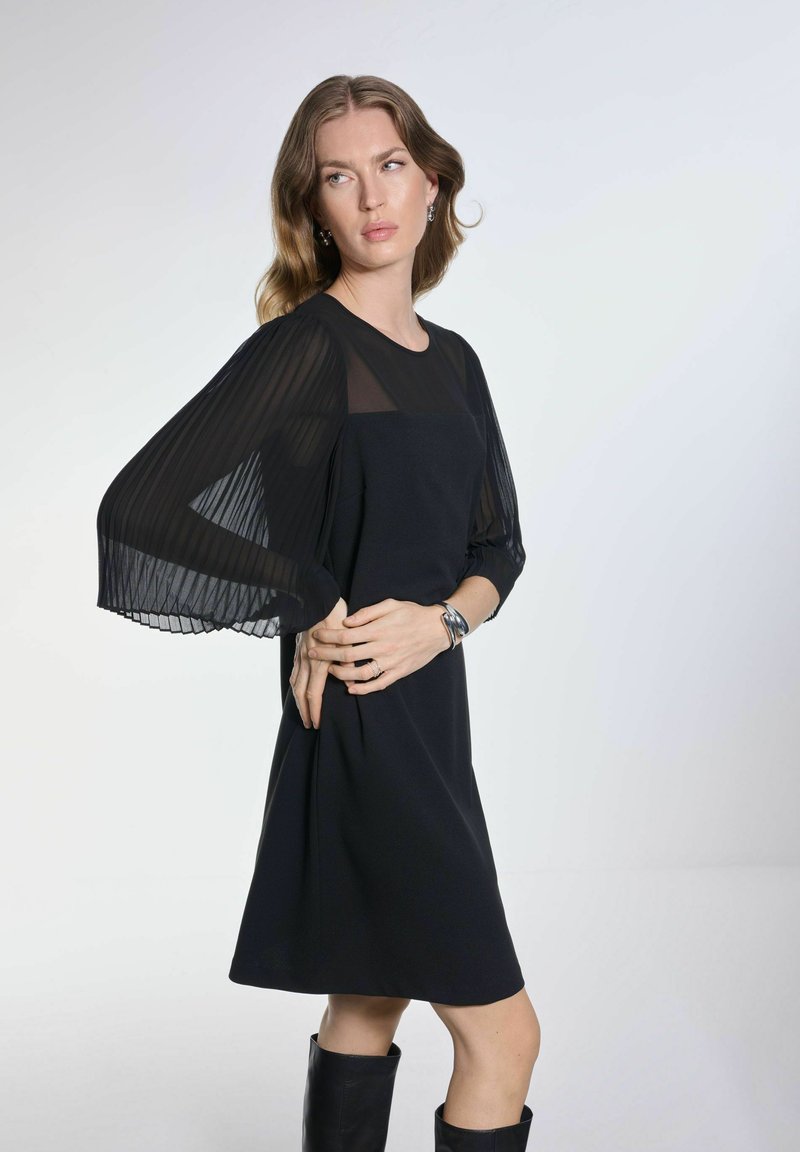 Czarna sukienka z przezroczystymi, plisowanymi długimi rękawami oraz dopasowanym korpusem. Gładka faktura z długością do kolan. Modelka nosi czarne buty za kolano.