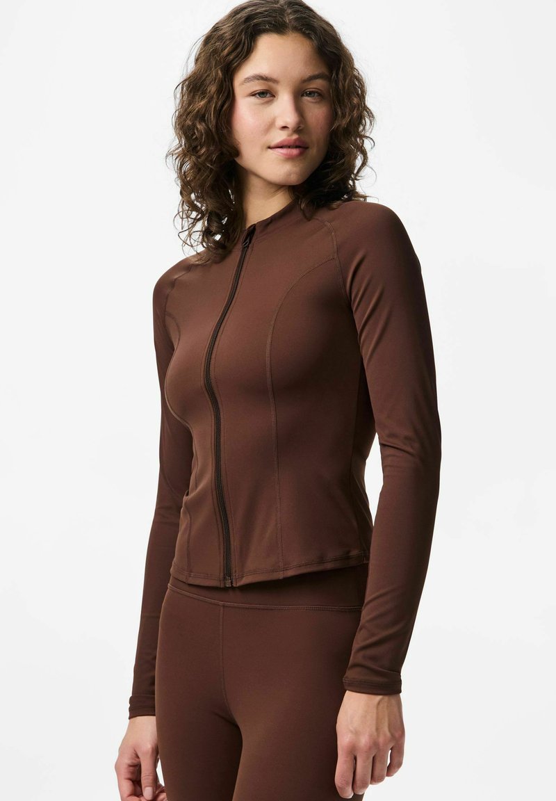 Rudos spalvos ilgomis rankovėmis sportinis užsegamas viršus, pagamintas iš tamprios medžiagos, pasižymintis glaudžiu siluetu su lygiais siūlėmis ir stovinčiu apykakliu.